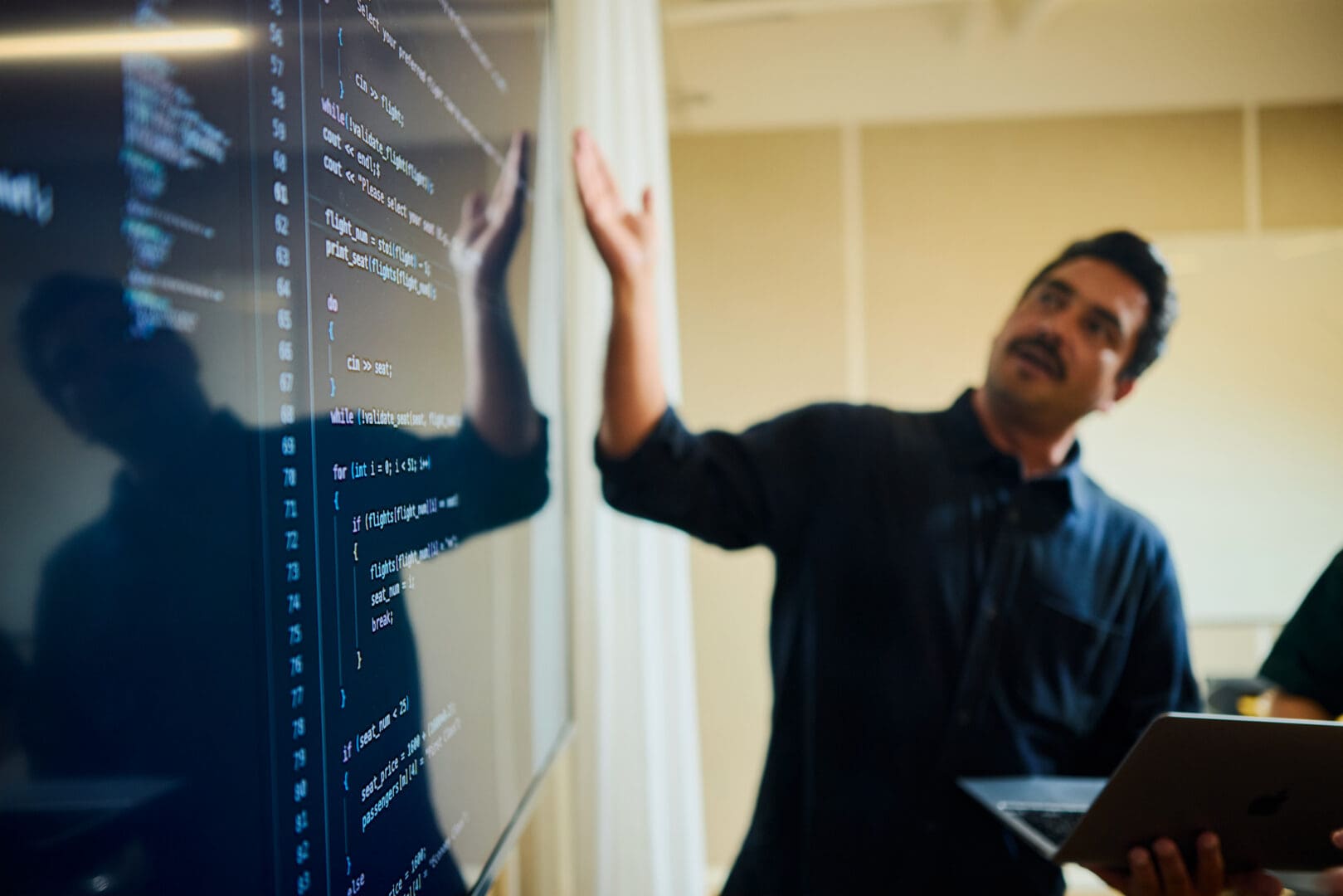 Woman pointing at code on a large screen while discussing with a colleague in an office